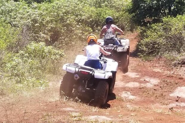 Two ATV riders following each other single file up a dusty red-dirt ranch track in the West Maui Mountains