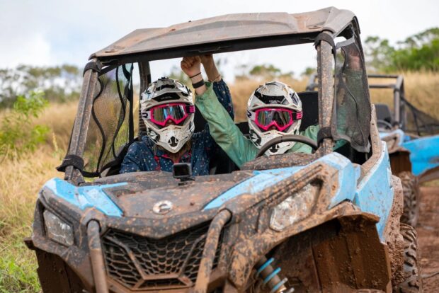 Two riders in helmets and goggles, mud-splattered, in a side-by-side ATV, one raising an arm