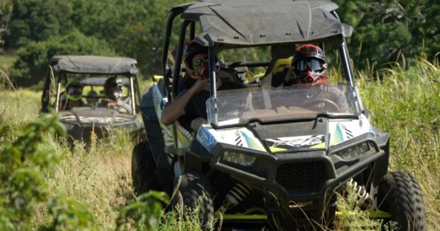 Two side-by-side ATVs with helmeted drivers moving through tall grass on a guided convoy