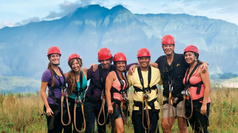 Group of seven guests in zipline harnesses posing together at Kipu Ranch, Haʻupu Mountain Range in the background