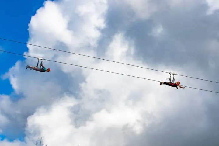Two guests fly side-by-side on the dual zipline at Nui Kauai in superman pose, with open sky behind them