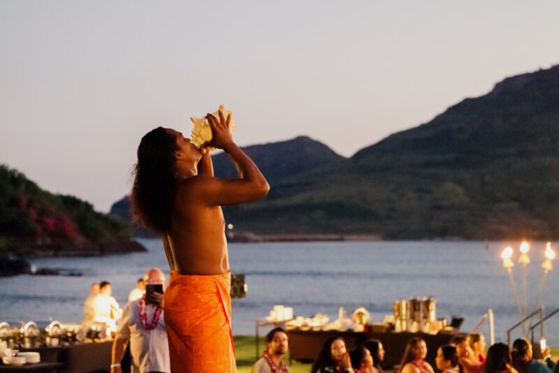 Performer blowing a conch shell at sunset during the Sonesta Kauaʻi Resort Luau, with Kalapaki Bay in the background.