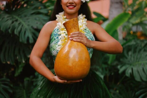 Hula dancer in a ti-leaf skirt and plumeria lei holding a large ipu heke gourd drum