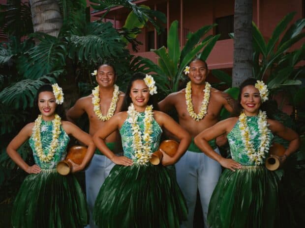 Five ʻAhaʻāina Luau dancers with ipu gourds posed in front of the pink walls of The Royal Hawaiian
