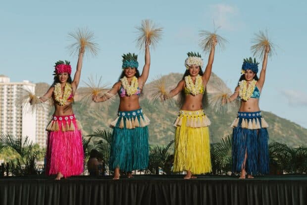 Four dancers in pink, teal, yellow and blue raffia skirts raising raffia tassels, Diamond Head behind