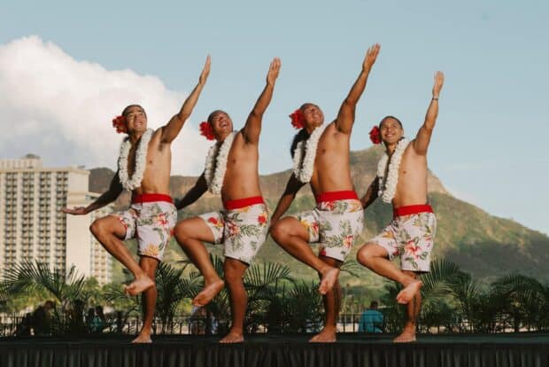 Four male hula dancers mid-step with one leg raised and arms extended, Diamond Head behind them