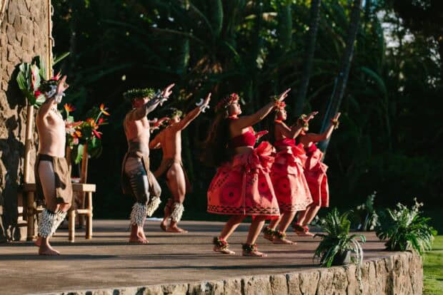 Hula dancers in red pāʻū skirts performing on stage at sunset at the Grand Wailea luau