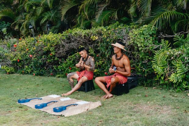 Two musicians in red kapa with ukuleles beside blue ukuleles set out for a lesson on the lawn