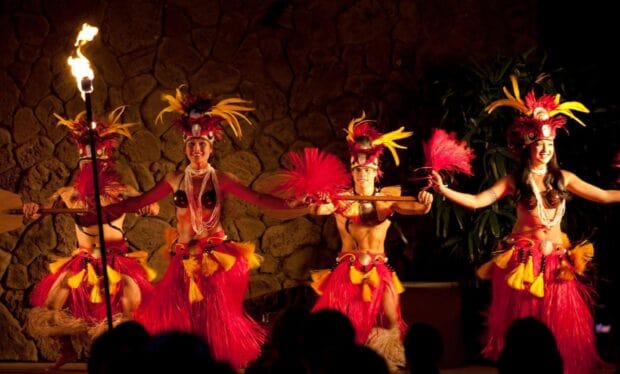 Dancers in red and yellow feather headdresses performing beside a lit fire torch at night