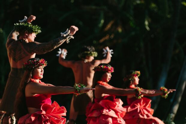 Close-up of hula dancers in coral pāʻū skirts and flower haku lei with arms extended at golden hour
