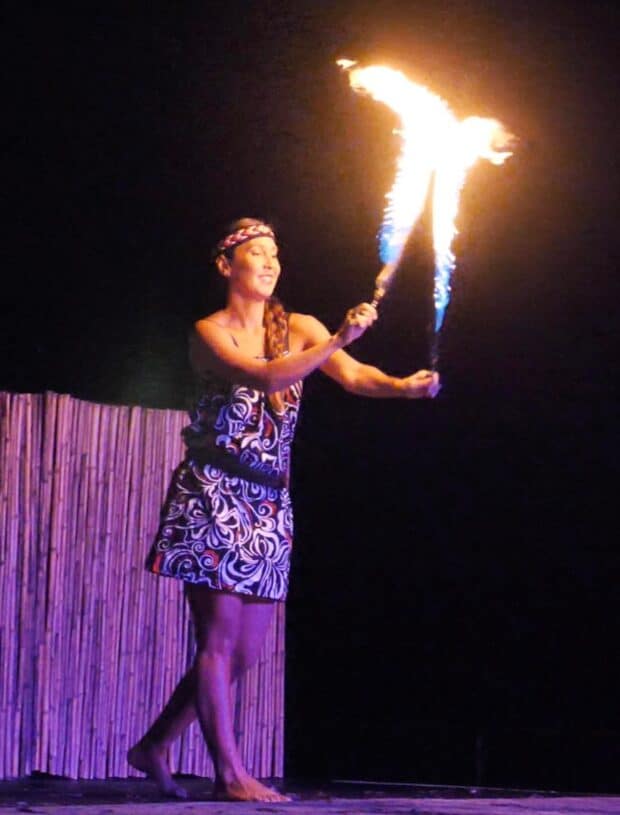 Performer in a patterned dress holds a flaming torch in both hands on the Ahi Lele Fire Show stage, Kauaʻi