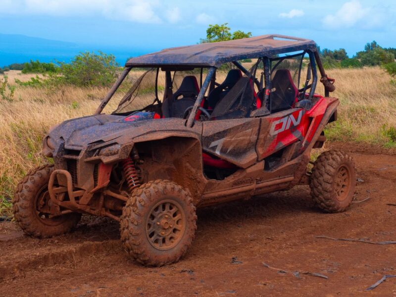 Four-seat side-by-side ATV caked in dried mud parked on a red-dirt clearing with hazy ocean behind