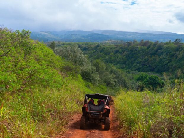 Four-seat side-by-side ATV on a red-dirt trail descending into a green valley with clouded West Maui mountains behind