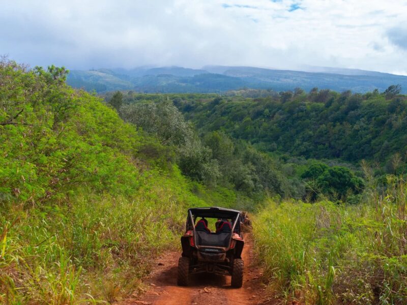 Four-seat side-by-side ATV on a red-dirt trail descending into a green valley with clouded West Maui mountains behind