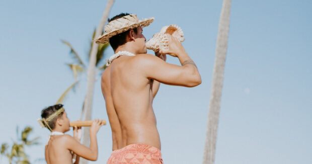 ʻAuliʻi Lūʻau performer in a woven hat blowing a conch shell under palm trees, a boy behind him