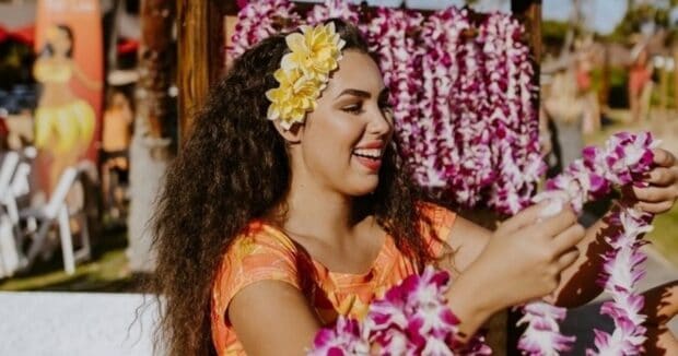 Smiling ʻAuliʻi Lūʻau staff member with yellow flowers in her hair holding purple and white orchid lei