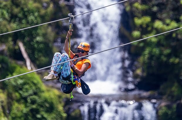 Smiling woman in orange helmet ziplining in front of a waterfall at the World Adventures Botanical Gardens, Big Island Hawaii