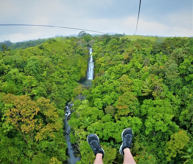 First-person view from the final zipline at Botanical World Adventures, feet dangling above Kamaʻeʻe Falls and rainforest gorge, Big Island Hawaii