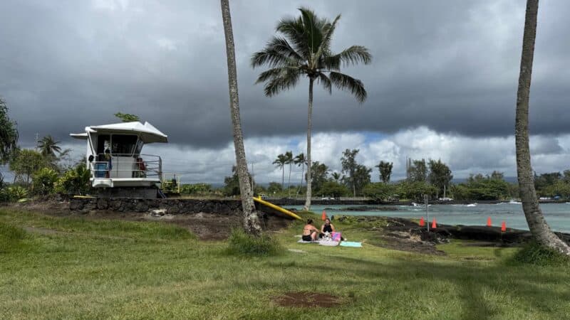 Lifeguard tower at Carlsmith Beach Park under heavy grey cloud, with two people on the lawn by the water