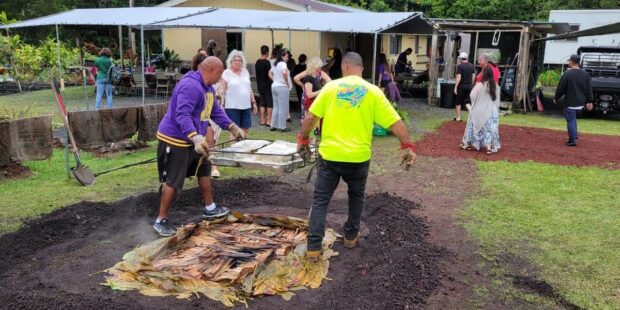 Two hosts carry a tray of cooked food from the opened imu across the yard at the Imu Mea ʻAi food tour