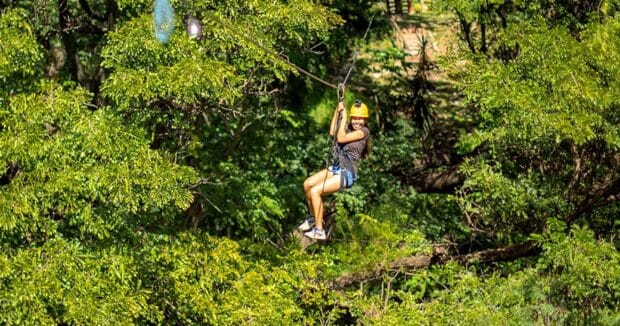 A rider crosses a long zipline span low over dense green treetops at Coral Crater, Oʻahu