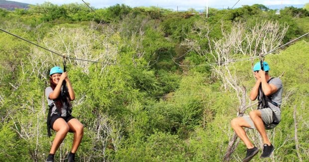 Two riders side by side on parallel zipline cables over green scrub forest at Coral Crater, Oʻahu