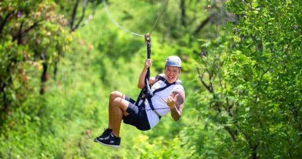 A man throws a shaka while ziplining through green trees at Coral Crater, Oʻahu