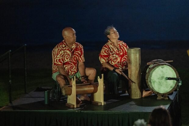 Two drummers in red print shirts play a wooden slit drum, a bamboo drum and a skin drum at the Royal Sonesta Kauaʻi lūʻau
