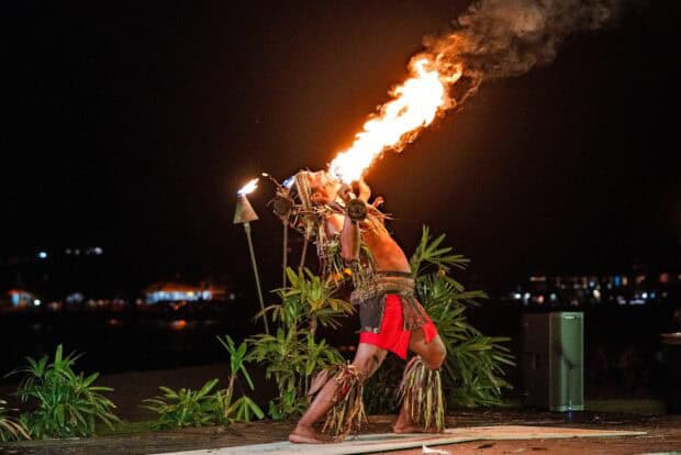 A fire dancer in a red wrap leans back with a burning staff on the Royal Sonesta Kauaʻi lūʻau stage