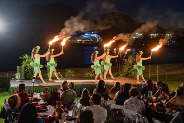 Five dancers twirl lit fire staffs on the Drums & Dances of Polynesia stage as lūʻau guests watch from tables on the lawn