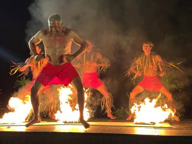 Male dancers in red wraps and leaf anklets dance between burning fire pots on stage at the Royal Sonesta Kauaʻi lūʻau