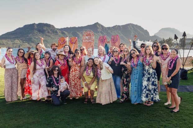 A group of guests in purple orchid lei poses on the lawn by Kalapaki Bay in front of carved Royal Sonesta Kauai signs