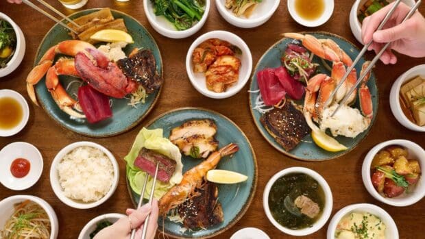 Plates of snow crab legs, sashimi, grilled fish and steak with banchan bowls on a wooden table