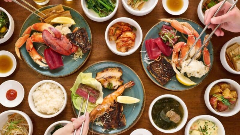 Plates of snow crab legs, sashimi, grilled fish and steak with banchan bowls on a wooden table