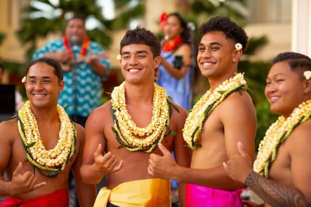 Four male hula dancers in stacked plumeria lei throw shaka in front of the musicians