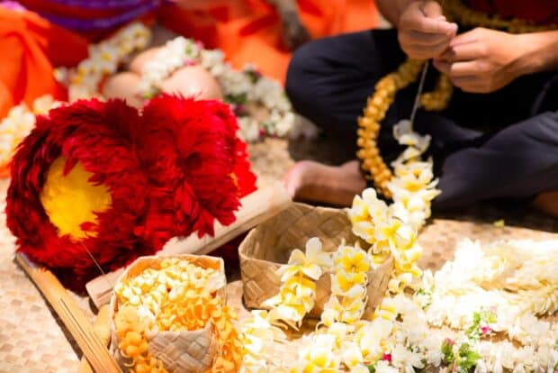 Hands stringing plumeria lei beside baskets of flowers and a red feather lei on a lauhala mat