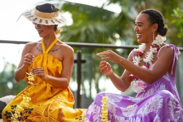 Two women in aloha wear laugh while stringing yellow plumeria lei on the terrace