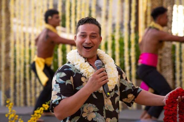 Host in a white carnation lei speaks into a microphone while dancers perform behind him