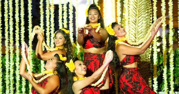 Five hula dancers in red pareo and yellow lei pose against the lei-strand backdrop