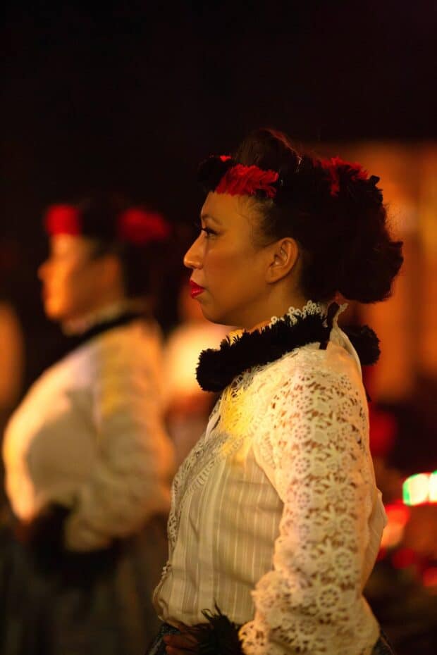 Performer in white lace with a red and black feather lei poʻo during the royal procession segment