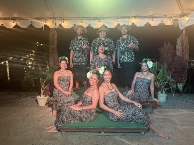 Eight cast members in matching leaf-print aloha wear pose under the tent at night