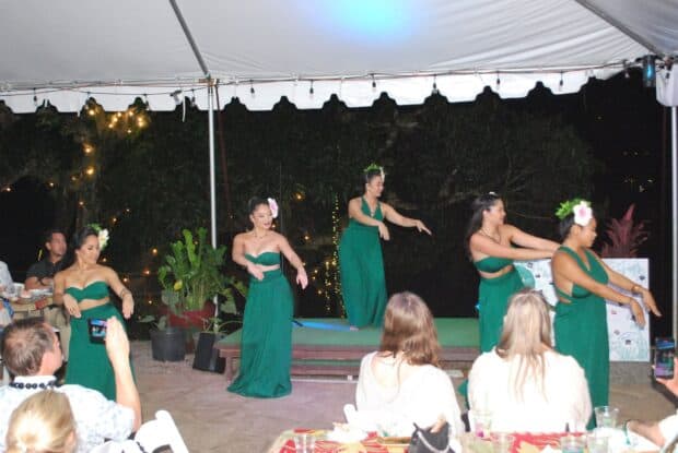 Five dancers in green dresses perform hula between guest tables under a white tent