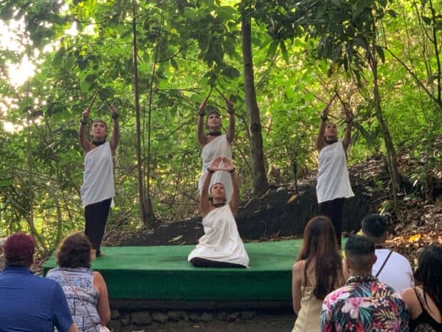 Four dancers in white kīhei raise kālaʻau sticks on a small stage in a forest clearing