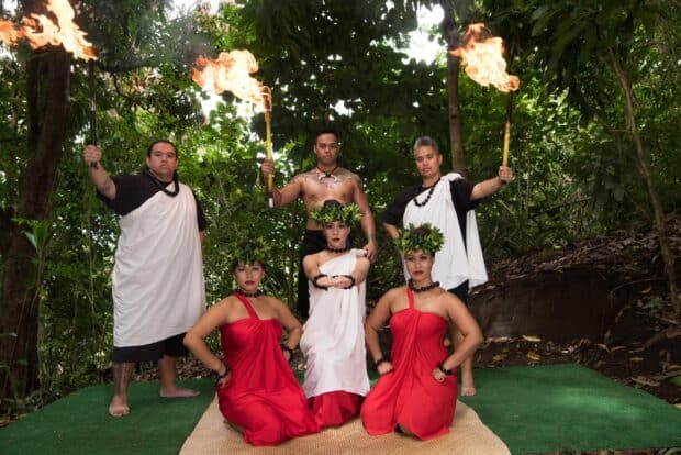 Six performers in red and white pose with burning torches in the forest
