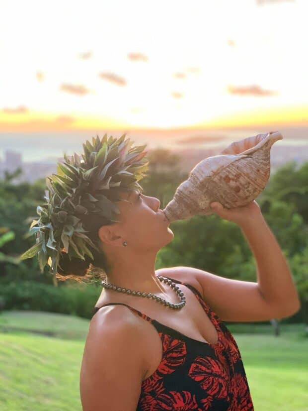 Performer in a lei poʻo blows a pū conch shell at sunset with Honolulu and the ocean far below