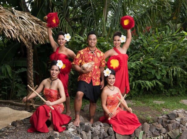 Five performers in red with ipu and pūʻili pose by a thatched-roof hale