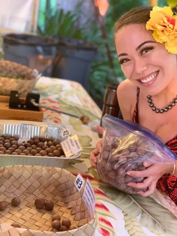 Performer holds a bag of macadamia nuts beside numbered trays of drying nuts and a cracking tool