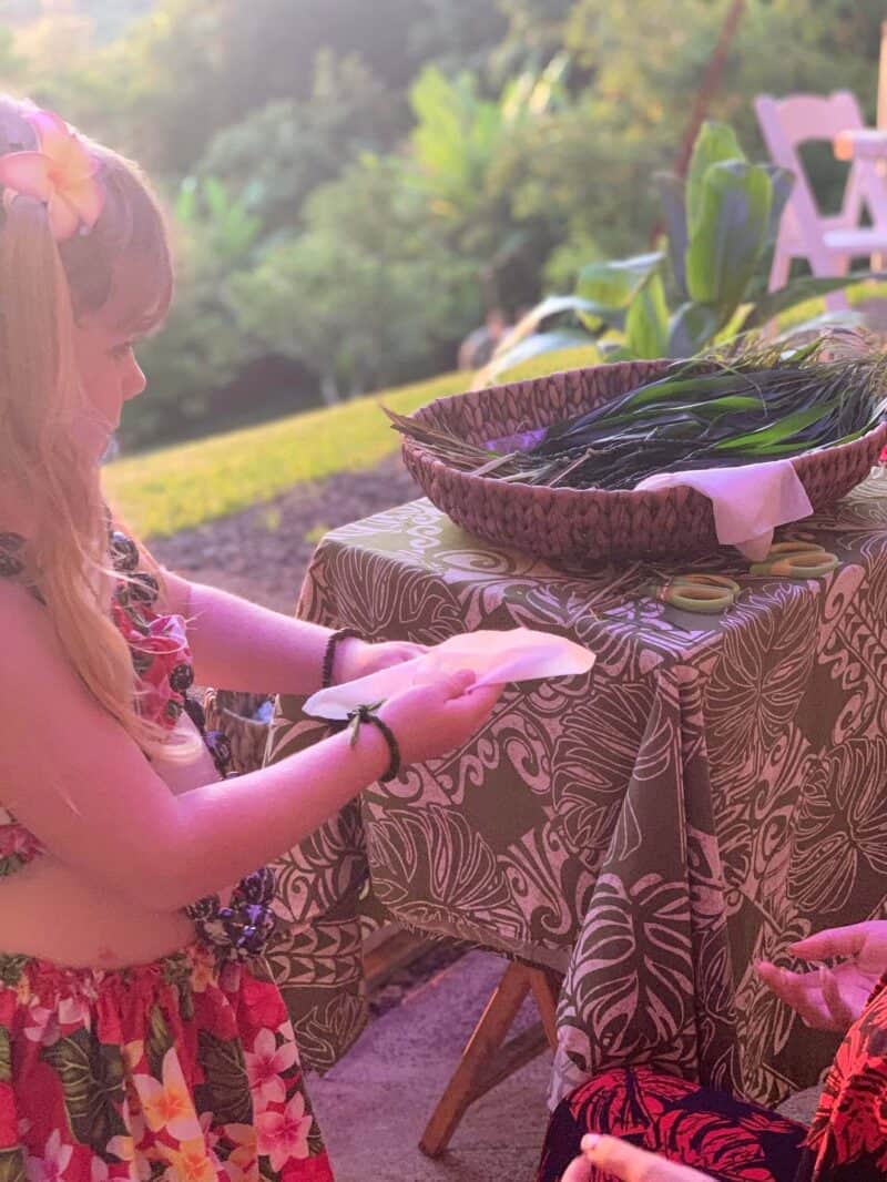 Young girl in a plumeria hairpiece strings a lei at a table on the lawn in late-day sun