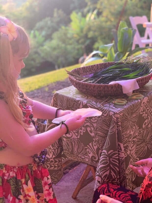 Young girl in a plumeria hairpiece strings a lei at a table on the lawn in late-day sun