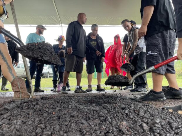 Guests shovel dark volcanic cinder over the imu under a tent in the rain at the Imu Mea ʻAi food tour